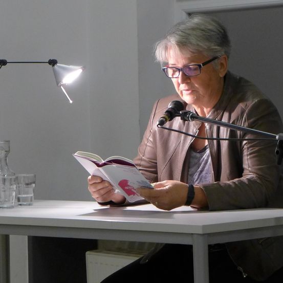 Eine ältere Frau liest von einem offenen Buch, während sie an einem Schreibtisch sitzt. Ein Mikrofon steht vor ihr. Zwei Lampen beleuchten den Bereich. Gläserne Behälter sind auf dem Schreibtisch.