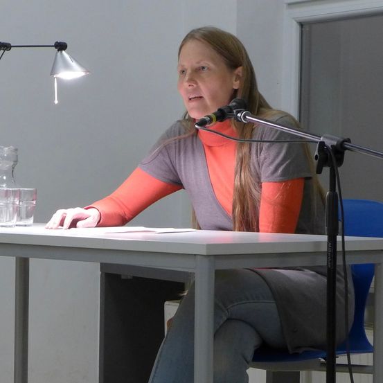 Eine Frau sitzt an einem Schreibtisch und spricht in ein Mikrofon. Sie trägt ein graues Rollkragenshirt mit orangefarbenen Ärmeln. Zwei Gläser und eine Flasche stehen auf dem Tisch. Eine Lampe und ein Heizkörper sind im Hintergrund zu sehen.