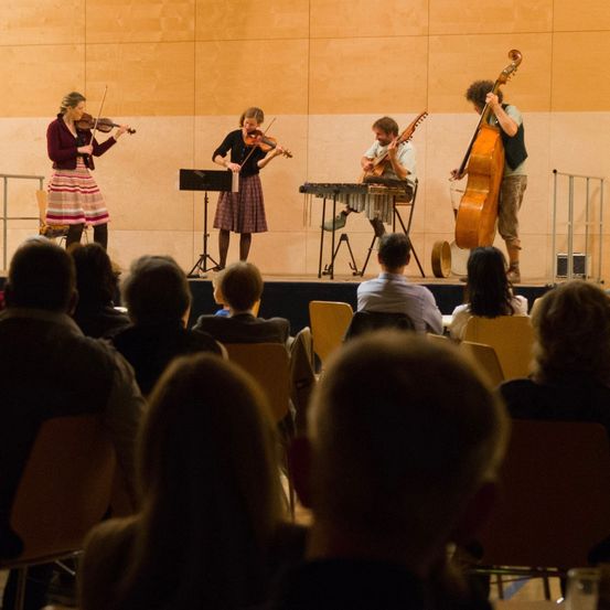 Drei Musiker spielen auf der Bühne, wobei zwei Frauen Violinen und ein Mann Cello spielen. Das Publikum sitzt vor ihnen auf Stühlen. Die Umgebung scheint ein Konzert zu sein.