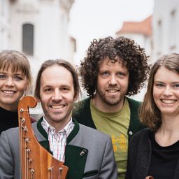 Vier Menschen lächeln und posieren für ein Foto. Zwei Frauen und zwei Männer. Der Mann in der Mitte hält eine Harfe. Dahinter steht ein Gebäude mit rotem Dach.