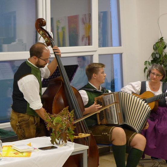 Ein Mann spielt einen großen Kontrabass, während ein anderer Akkordeon spielt. Sie befinden sich in einem Raum mit großen Fenstern. Auf einem Tisch vor ihnen stehen ein Buch, ein Telefon, eine Kanne und ein Glas.