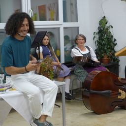 Ein Mann mit lockigem Haar sitzt und hält ein Tablet in der Hand, lächelt. Neben ihm steht ein Cello. Eine Frau sitzt auf einem Stuhl mit einer Gitarre. Eine weitere Frau sitzt hinter ihnen. Es gibt eine Pflanze und einen Tisch mit Büchern.