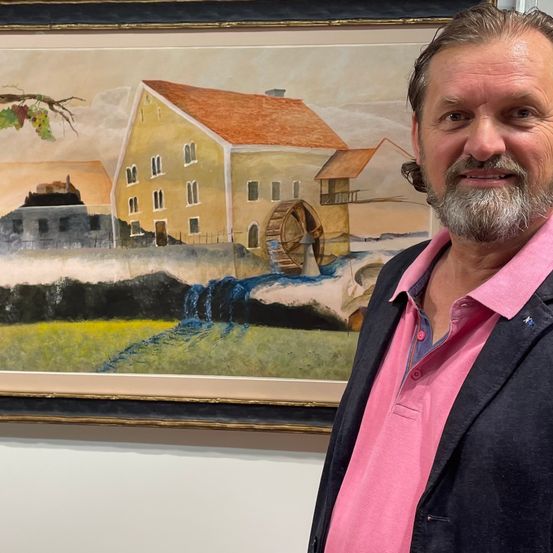 Ein Mann in einem rosa Hemd und einem dunklen Blazer steht vor einem gerahmten Gemälde, das eine ländliche Landschaft mit einer Wassermühle darstellt.