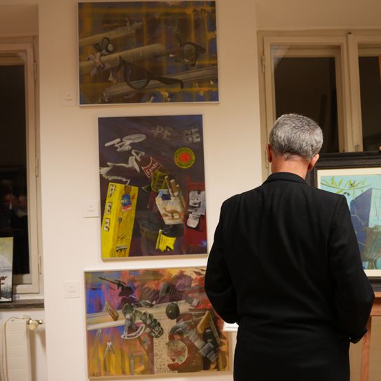 Ein Mann in einem schwarzen Anzug betrachtet Kunstwerke an einer weißen Wand mit drei Gemälden. Die Gemälde enthalten abstrakte Designs und Collagen mit verschiedenen Objekten.