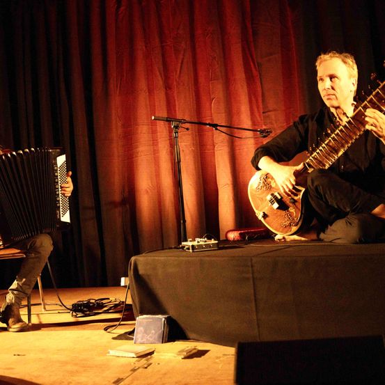 Zwei Musiker spielen auf der Bühne. Einer spielt Akkordeon und der andere Sitar. Ein Mikrofon steht vor ihnen.