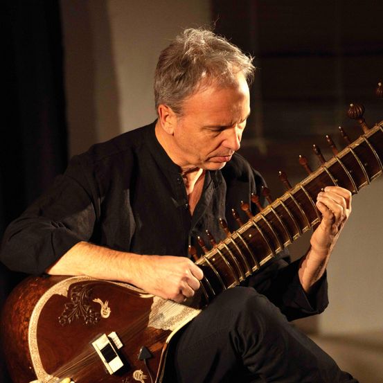 Ein Mann in einem schwarzen Hemd spielt ein Sitar, ein traditionelles indisches Saiteninstrument, in einem schwach beleuchteten Raum.