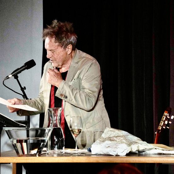 Ein älterer Mann steht an einem Podium, hält ein Papier in der Hand und spricht in ein Mikrofon. Auf einem Tisch davor befinden sich eine Gitarre, ein Weinglas und eine Karaffe.