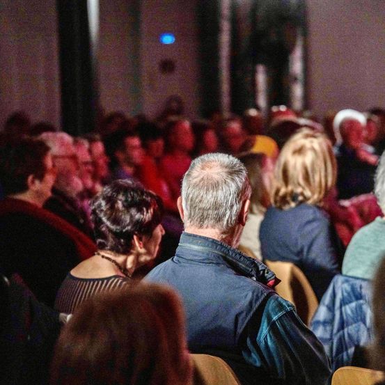 Ein großes Publikum sitzt in einem schwach beleuchteten Raum, nach vorne gerichtet. Einige Leute tragen Hüte und Brillen. Der Raum ist mit Menschen verschiedenen Alters gefüllt.