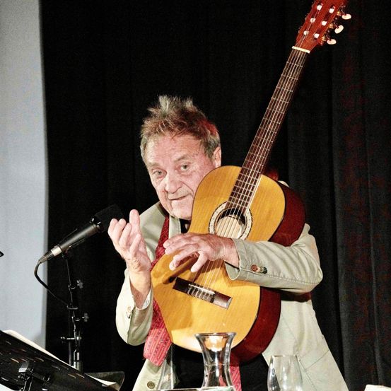 Ein älterer Mann steht auf der Bühne mit einer Gitarre, lächelt und gestikuliert, während er das Instrument hält. Ein Mikrofon ist vor ihm positioniert.