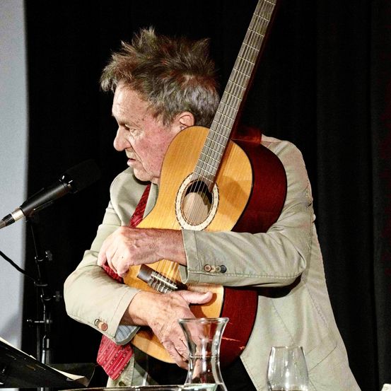 Ein Mann auf der Bühne, der eine Gitarre hält, möglicherweise bei einer Aufführung, mit Mikrofon, schwarzem Hintergrund und Weinglas.