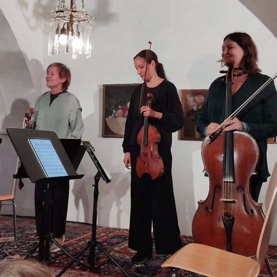 Two women are playing violins in a room with a chandelier, a painting, and a carpet. A music stand and a chair are nearby. A man plays a clarinet.