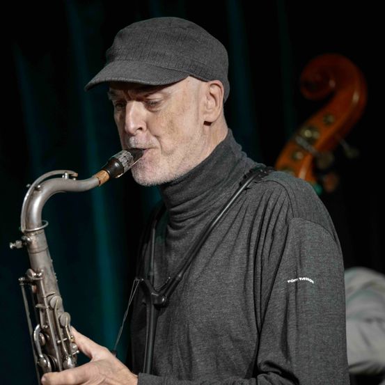 An older man plays a saxophone on stage. He wears a black hat and a gray turtleneck shirt. A cello is visible behind him.