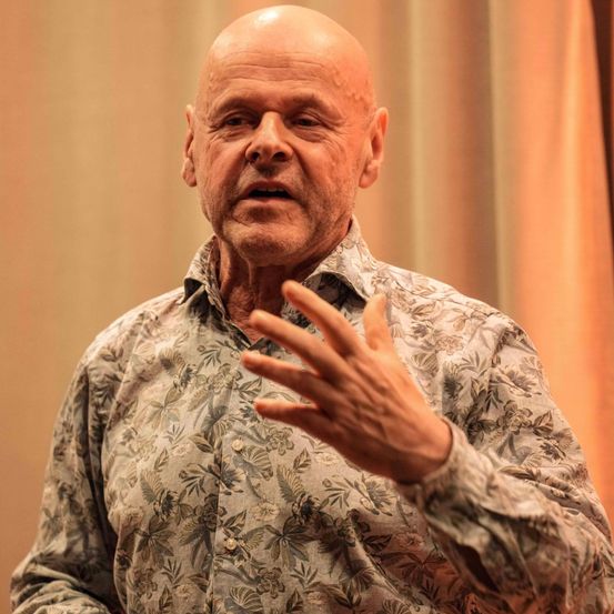 A bald man in a floral shirt stands and speaks, gesturing with his hands. He looks serious and focused.