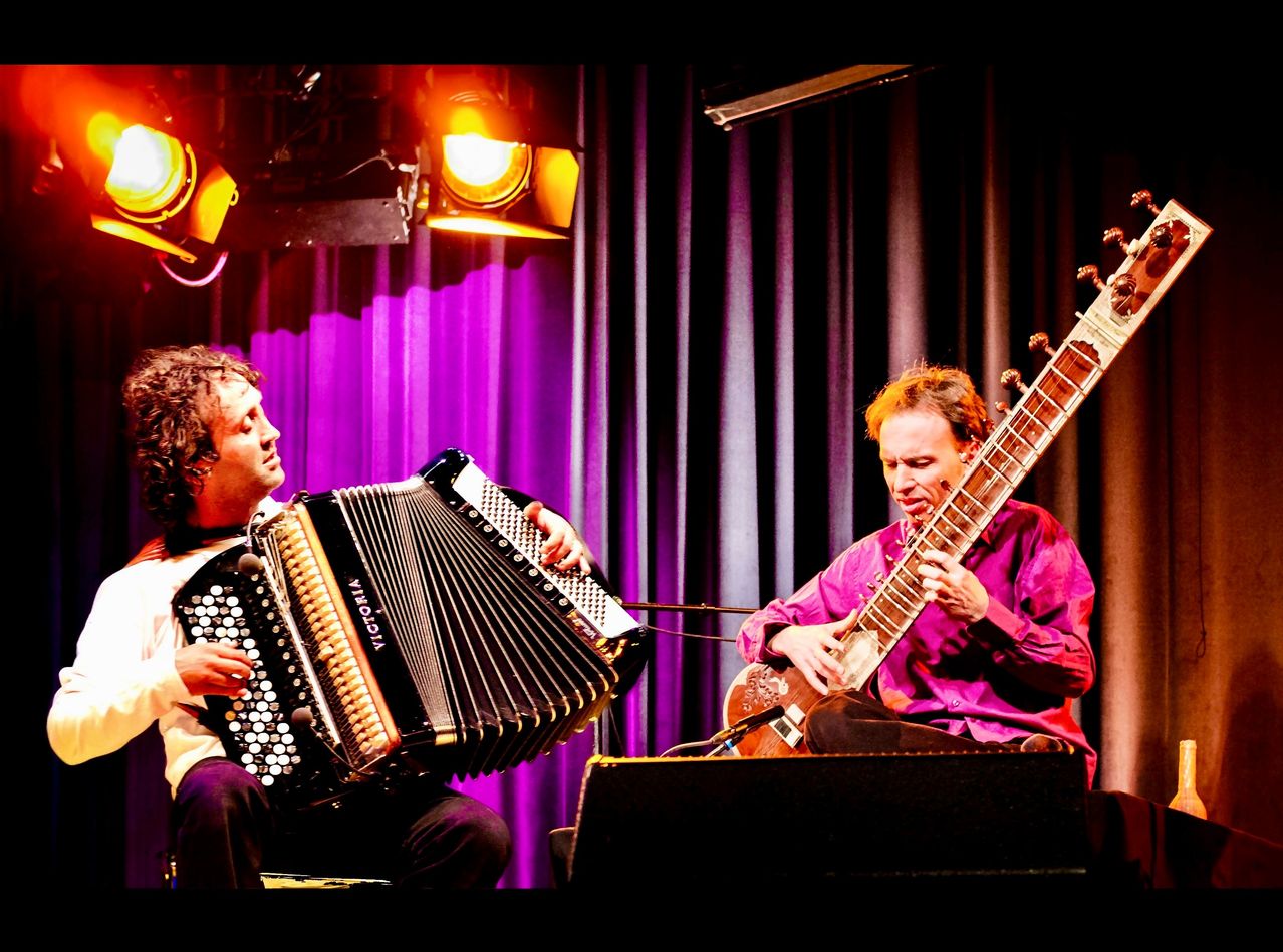 Two musicians perform on stage under spotlights. One plays an accordion, the other a sitar. Both wear purple long-sleeved shirts.
