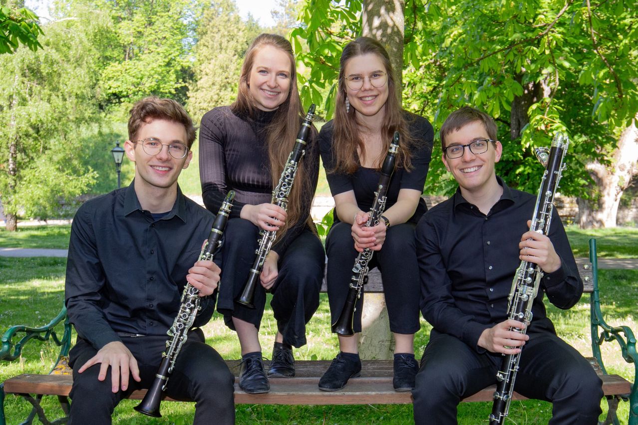 Fünf Musiker in schwarzer Kleidung posieren mit ihren Klarinetten in einem Park. Zwei Frauen in der Mitte lächeln, mit Bäumen im Hintergrund.