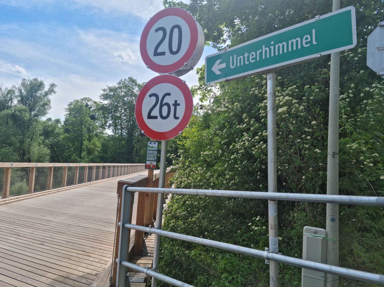 Eine Brücke mit zwei Verkehrsschildern: 20 km/h Geschwindigkeitsbegrenzung und 26 Tonnen Gewichtsbegrenzung. Grünes Richtungsschild für Unterhimmel.