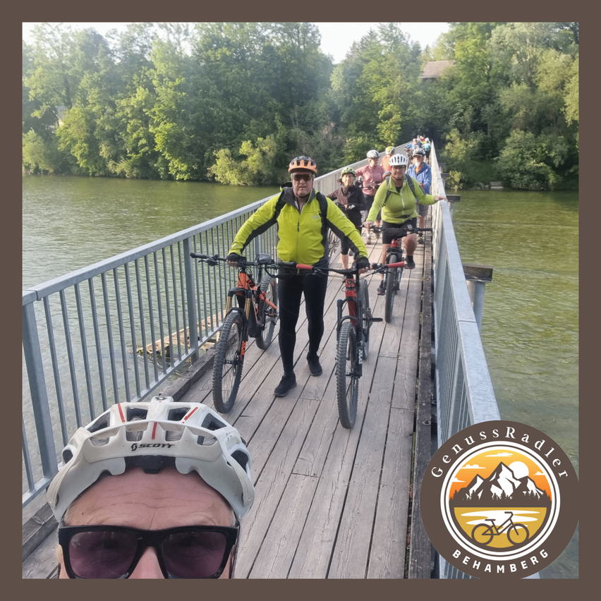 Eine Gruppe von Radfahrern mit Helmen und Radtrikots steht auf einer Holzbrücke über einem Fluss, mit grünem Wasser und Bäumen im Hintergrund.