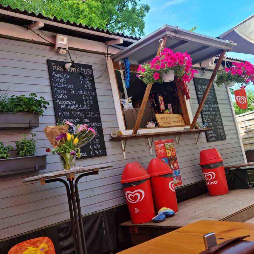 Ein Imbissstand mit Kreidetafel-Menü und rosa Blumen. Rote Mülltonnen und ein kleiner Tisch mit Blumen. Der Stand befindet sich neben einer Holzterrasse.