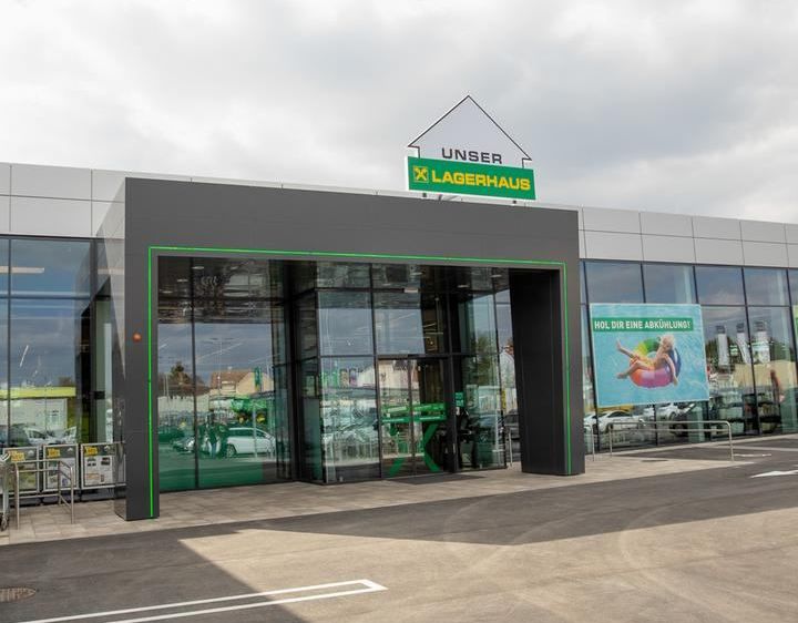 Der Eingangsbereich eines großen Gebäudes zeigt eine Glastür mit grünem Rand und einem Schild, das 'Unser Lagerhaus' besagt. Das Gebäude ist modern mit einer grauen Fassade und Glasfenstern. Auf der rechten Seite des Gebäudes befindet sich ein Plakat.