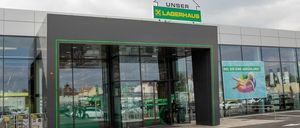 Der Eingangsbereich eines großen Gebäudes zeigt eine Glastür mit grünem Rand und einem Schild, das 'Unser Lagerhaus' besagt. Das Gebäude ist modern mit einer grauen Fassade und Glasfenstern. Auf der rechten Seite des Gebäudes befindet sich ein Plakat.