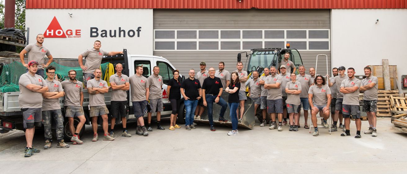 Eine Gruppe von Arbeitern, die graue Uniformen tragen, steht vor einem Gebäude mit roter Außenfassade. Ein Arbeiter sitzt auf einem Lastwagen, während andere vor einem Baufahrzeug stehen.