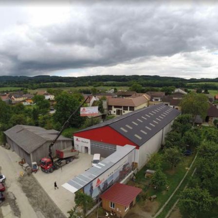 Luftaufnahme einer Baustelle mit Gebäuden, Kränen, geparkten Autos und einer hügeligen Landschaft.