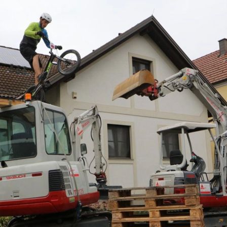 Eine Person fährt mit einem Fahrrad auf einem rot-weißen Bagger vor einem Haus mit Solarmodulen.
