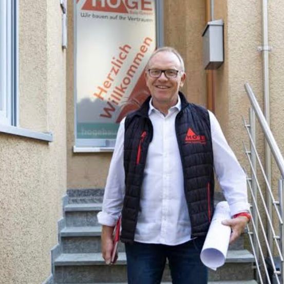 Ein Mann mit Brille und einem schwarzen Weste steht auf den Stufen eines Gebäudes, hält ein aufgerolltes Papier in der Hand und lächelt. Hinter ihm befindet sich ein Schild mit den Worten 'Herzlich Willkommen' und anderem Text.