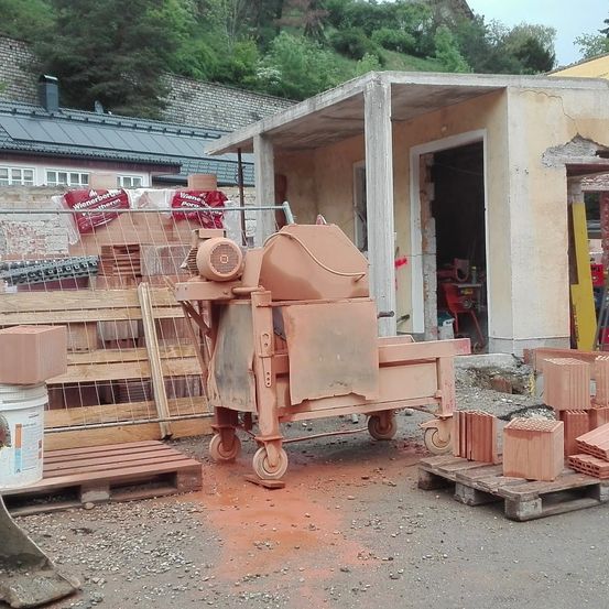 Eine Baustelle mit einer Ziegelherstellungsmaschine, einem Haufen Ziegel und einem Gebäude in Renovierung. Ein Holzpalette hält einen Eimer mit einem Etikett.