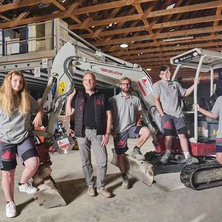 Fünf Personen stehen neben einem Takeuchi-Bagger in einem Lagerhaus. Sie sind in Arbeitskleidung gekleidet und scheinen für ein Foto zu posieren.