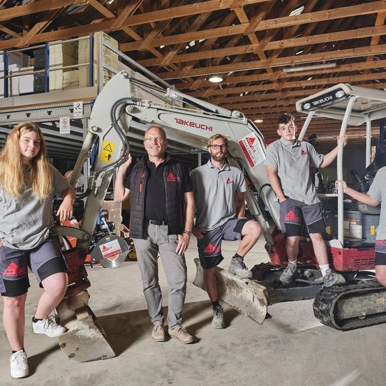 Fünf Personen stehen neben einem Takeuchi-Bagger in einem Lagerhaus. Sie sind in Arbeitskleidung gekleidet und scheinen für ein Foto zu posieren.