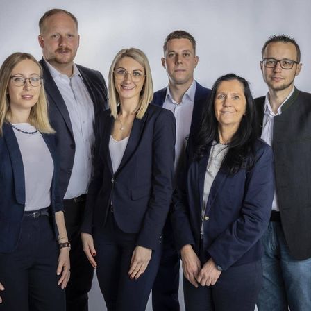 Eine Gruppe von sechs Fachleuten, drei Männer und drei Frauen, gekleidet in Geschäftskleidung, posiert für ein Foto im Studio. Sie stehen eng beieinander, lächeln und scheinen Kollegen zu sein.
