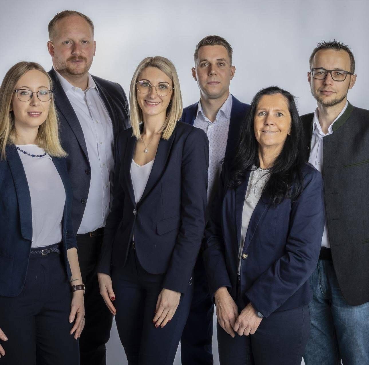 Eine Gruppe von sechs Fachleuten, drei Männer und drei Frauen, gekleidet in Geschäftskleidung, posiert für ein Foto im Studio. Sie stehen eng beieinander, lächeln und scheinen Kollegen zu sein.