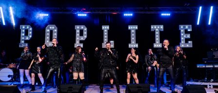 Eine Gruppe von Künstlern in schwarzen Outfits steht auf der Bühne, singt in Mikrofone und hat blaue Lichter und 'OPLIT' in leuchtenden Buchstaben im Hintergrund.