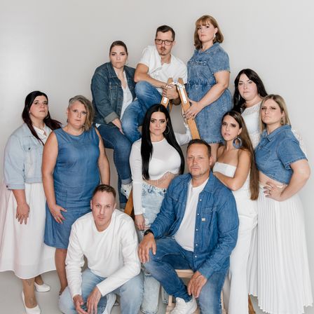 Eine Gruppe von Erwachsenen, sowohl Männer als auch Frauen, posiert für ein Foto im Studio. Einige stehen, während andere auf einer Leiter oder einem Stuhl sitzen. Sie sind alle lässig gekleidet.