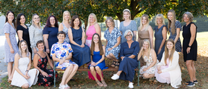 Eine Gruppe von Frauen in verschiedenen Kleidern posiert für ein Foto unter einem Baum in einem Park. Sie sitzen auf einer Bank und stehen in einer Reihe.