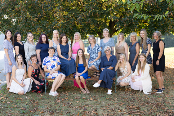 Eine Gruppe von Frauen in verschiedenen Kleidern posiert für ein Foto unter einem Baum in einem Park. Sie sitzen auf einer Bank und stehen in einer Reihe.
