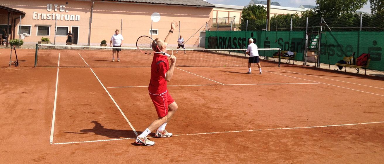 Drei Männer spielen Tennis auf einem Sandplatz. Ein Mann in Rot bereitet sich darauf vor, den Ball zu schlagen, während ein anderer Mann in Weiß sich dem Netz nähert. Ein gelber Ball ist in der Luft.
