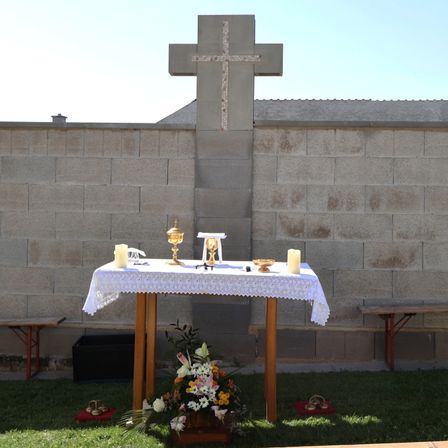 Ein einfacher Altaraufbau mit einem Kreuz an einer Steinmauer, einem Tisch mit einer weißen Tischdecke und Blumenarrangements. Zwei Kerzen stehen auf dem Tisch, neben einem Kelch und einer Schale.