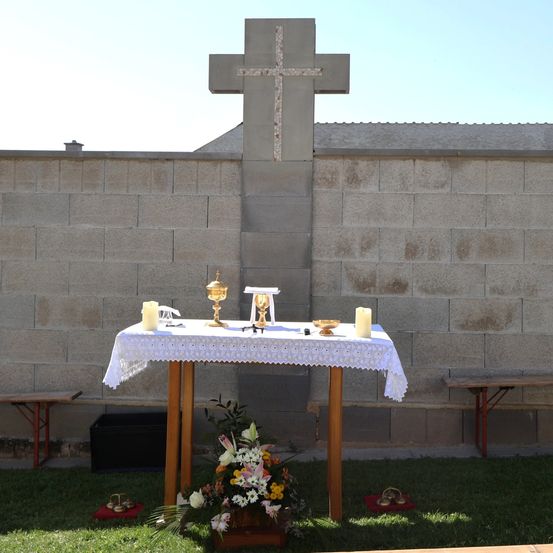 Ein einfacher Altaraufbau mit einem Kreuz an einer Steinmauer, einem Tisch mit einer weißen Tischdecke und Blumenarrangements. Zwei Kerzen stehen auf dem Tisch, neben einem Kelch und einer Schale.