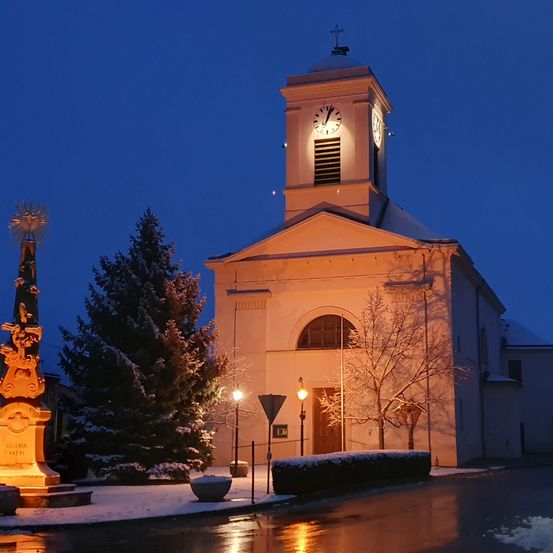 Eine Kirche mit Turm und Uhr steht in der Nacht beleuchtet, umgeben von Schnee und Bäumen.