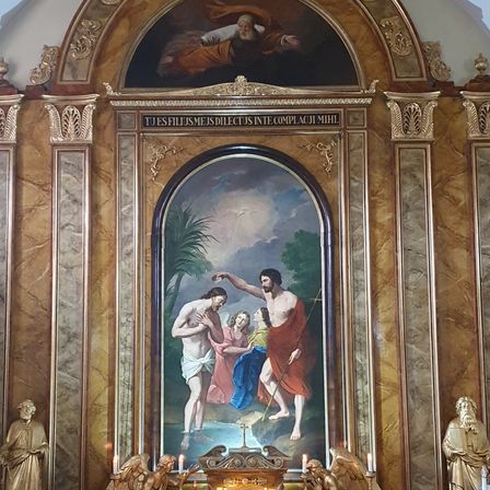 Ein religiöses Gemälde zeigt Jesus, wie er einen Mann in einem Fluss mit zwei Frauen tauft. Über dem Gemälde steht eine lateinische Phrase. Das Gemälde wird von goldenen Säulen und Statuen auf beiden Seiten gerahmt.