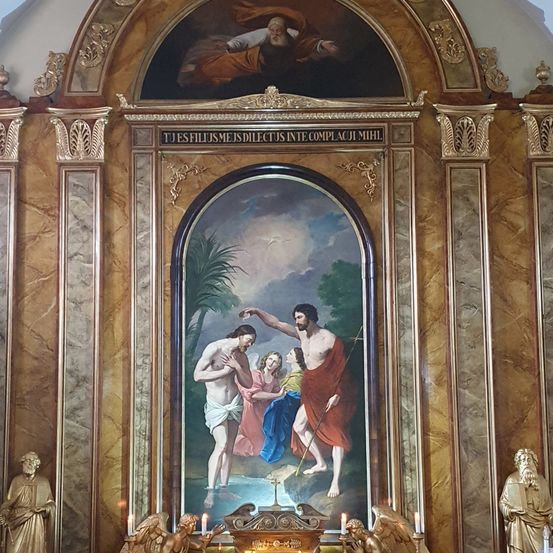 Ein religiöses Gemälde zeigt Jesus, wie er einen Mann in einem Fluss mit zwei Frauen tauft. Über dem Gemälde steht eine lateinische Phrase. Das Gemälde wird von goldenen Säulen und Statuen auf beiden Seiten gerahmt.