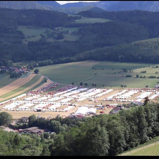 Eine Luftaufnahme eines großen Freiluftevents mit zahlreichen Zelten in einem Feld, umgeben von Bäumen und Hügeln in der Ferne.