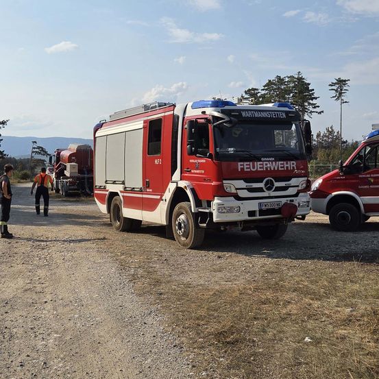 Ein roter Mercedes-Benz Feuerwehrwagen steht auf einem Schotterweg, zwei Personen in orangefarbenen Westen stehen in der Nähe. Ein weiteres Fahrzeug ist dahinter geparkt.