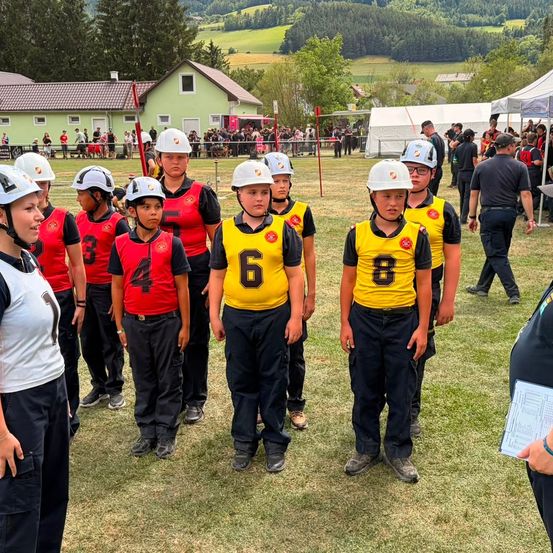 Eine Gruppe junger Menschen in gelben und roten Uniformen mit Nummern auf dem Rücken steht auf einem Feld, wahrscheinlich für eine Sportveranstaltung. Sie tragen Helme und Turnschuhe. Hinter ihnen befinden sich Zelte, eine Menschenmenge und ein Gebäude.