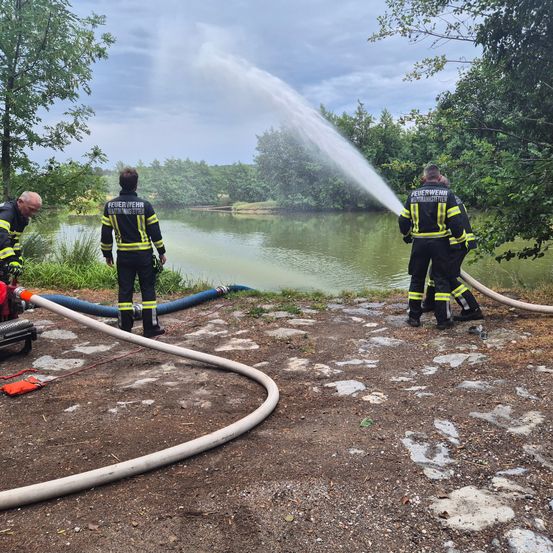 Drei Feuerwehrleute in Uniform sprühen Wasser aus einem Schlauch in einen See. Die Szene ist im Freien mit Bäumen und einem bewölkten Himmel.