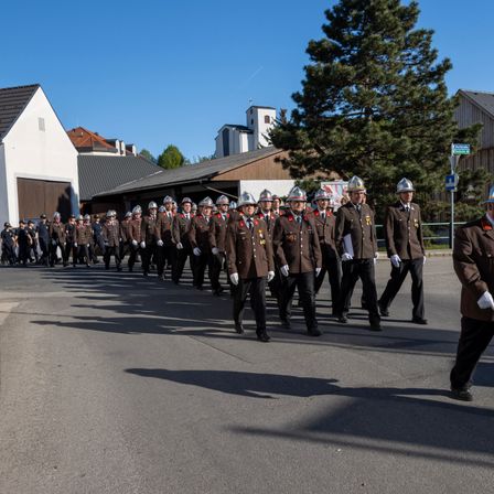 Eine Reihe uniformierter Personen, möglicherweise Feuerwehrleute, marschiert bei einem Umzug an einem sonnigen Tag.