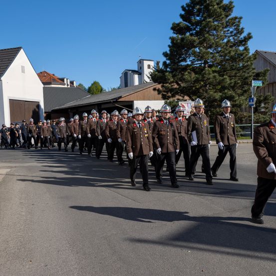 Eine Reihe uniformierter Personen, möglicherweise Feuerwehrleute, marschiert bei einem Umzug an einem sonnigen Tag.