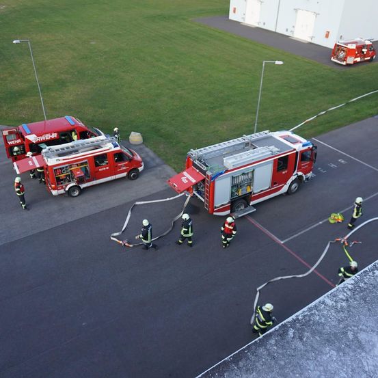 Eine Vogelperspektive von Feuerwehrleuten in voller Ausrüstung, die einen Feuerwehrwagen bedienen und Schläuche auslegen auf einem gepflasterten Bereich. Ein zweiter Feuerwehrwagen ist geparkt in der Nähe, und ein dritter ist im Hintergrund sichtbar.